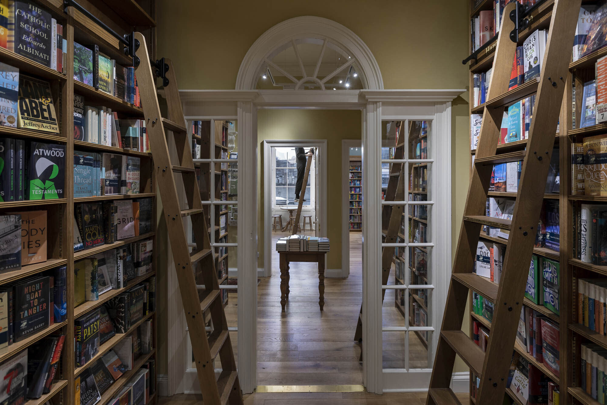 Library ladders to reach high shelves in the Topping and Company bookshop, in Edinburgh's Georgian New Town. A doorway with a fan light leads through to further booklined rooms, and a member of staff climbs a ladder.